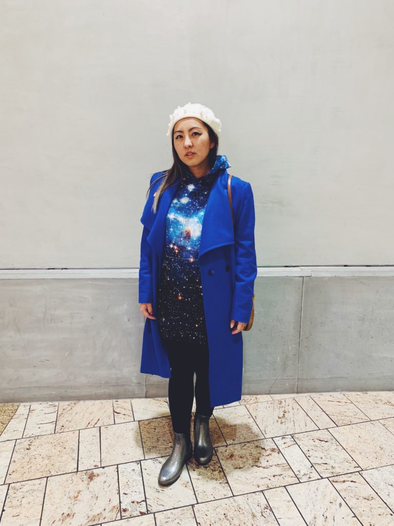 Alice wearing a long, blue galaxy-print hoodie under a long blue coat with black leggings and silver boots.