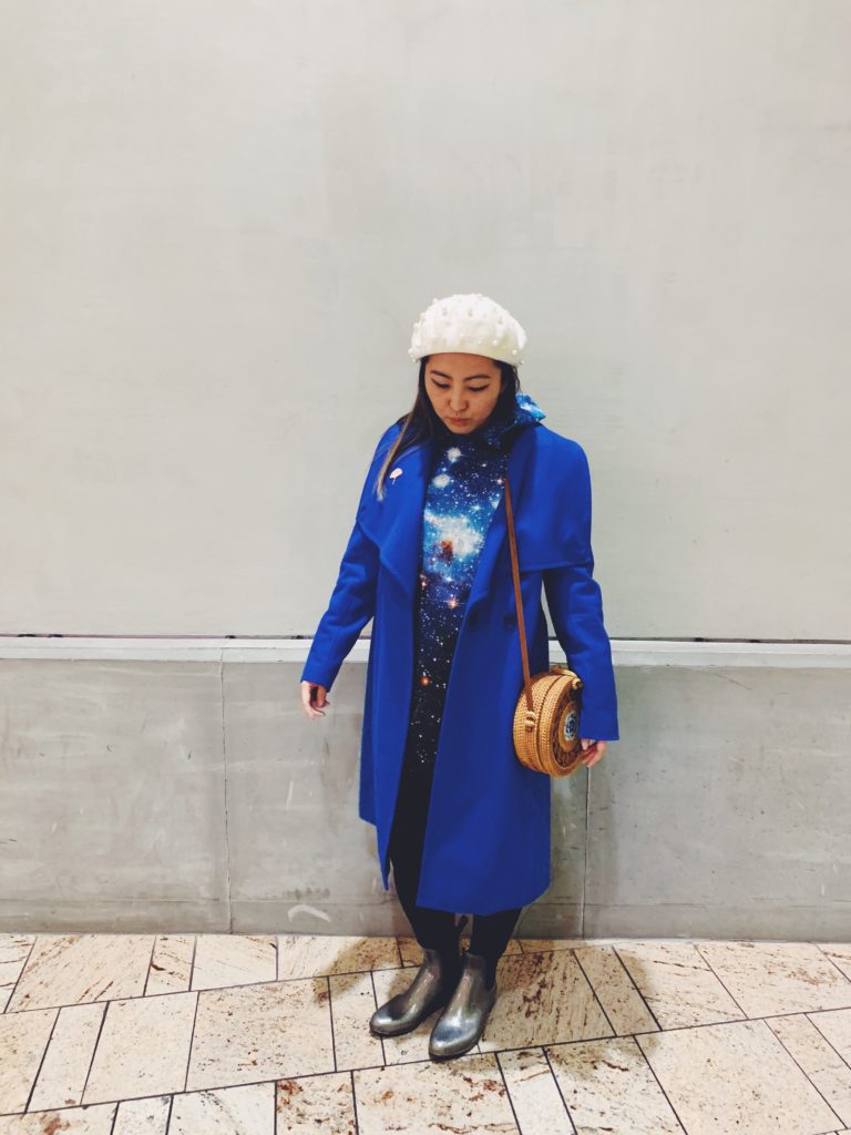Alice wearing a white beret, a blue galaxy-print hoodie under a long blue coat with black leggings and silver boots.