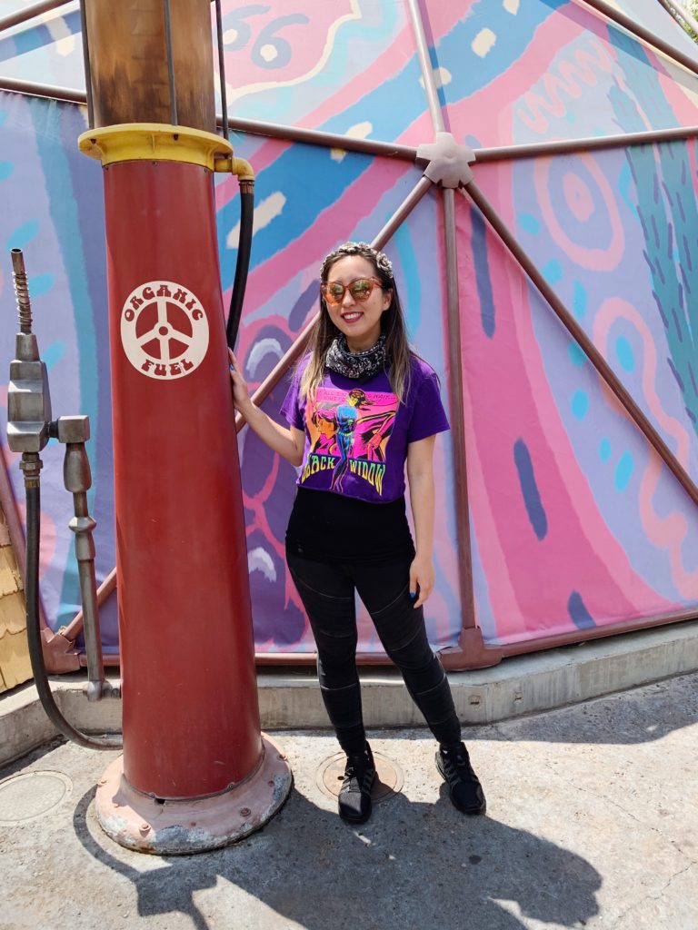 Festival-ready outfit featuring a purpled cropped Black Widow T-Shirt. Background: a pink and blue tent and a fake fueling pump.