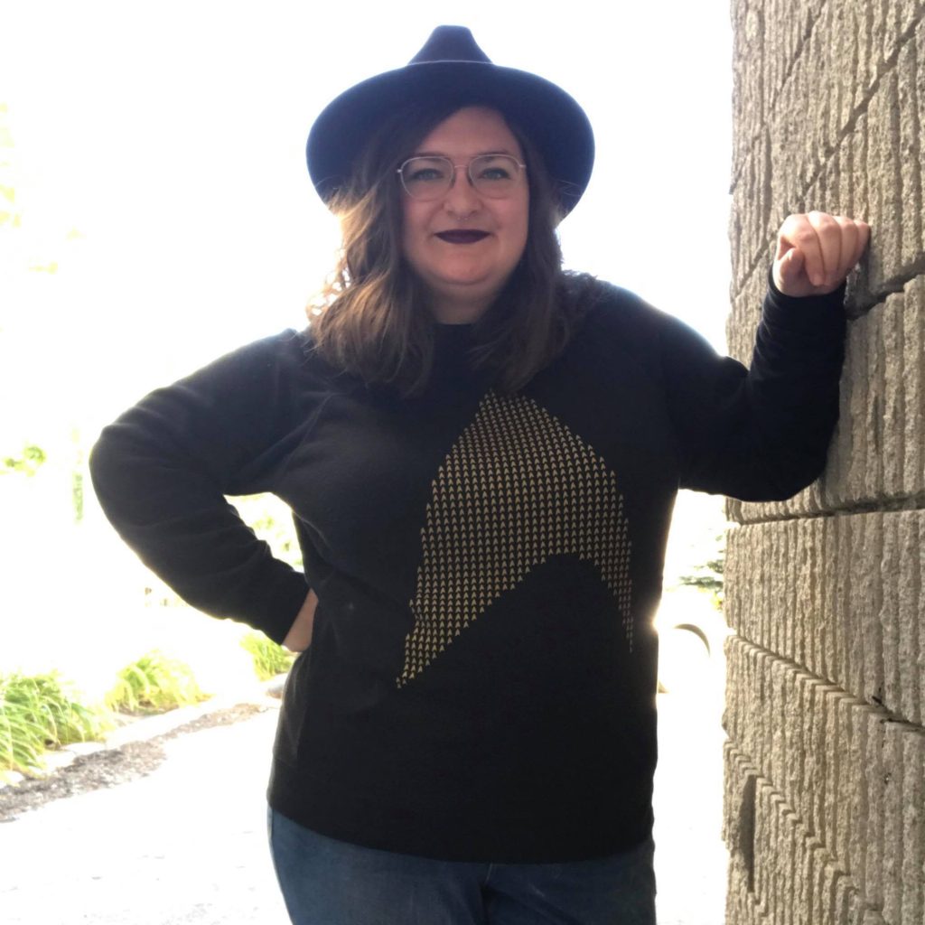 Brittany is leaning against a brick wall, smiling at the camera. They are wearing a sweatshirt with a golden star trek logo, and a black fedora.