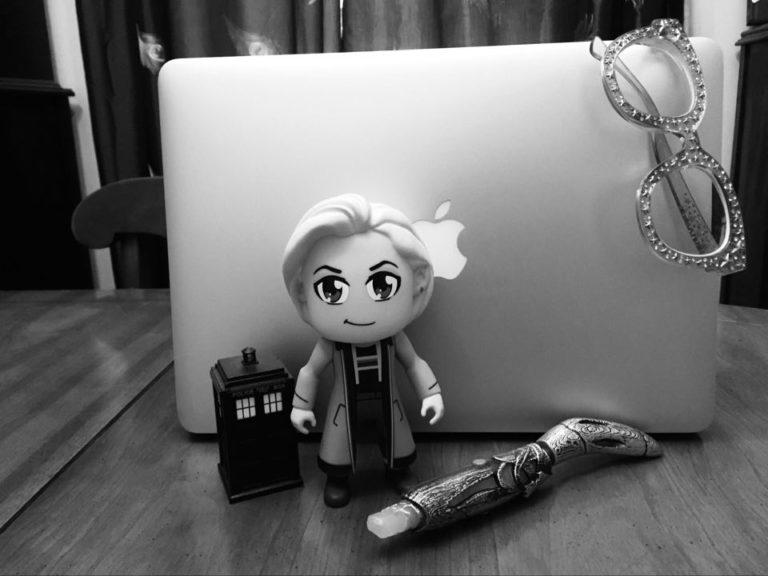 A Macbook is surrounded by a tiny TARDIS figure, a 13th Doctor figurine, a 13th Doctor sonic screwdriver (from Doctor Who) and a pair of star-speckled glasses hang off the right hand corner of the Macbook.